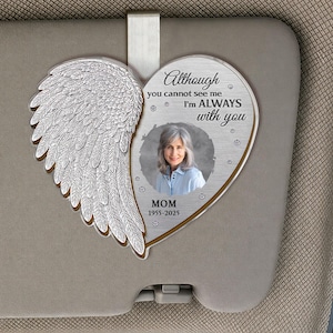 Puede incluir: Un adorno conmemorativo en forma de corazón con un ala de ángel plateada a la izquierda y una foto de una mujer a la derecha. El texto dice "Aunque no puedes verme, SIEMPRE estoy contigo" y "MAMÁ 1955-2025". El adorno cuelga de un parasol de coche.