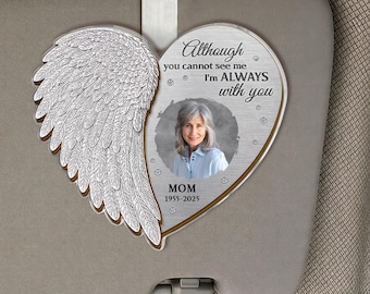 May include: A heart-shaped memorial ornament with a silver angel wing on the left and a photo of a woman on the right. The text reads "Although you cannot see me, I'm ALWAYS with you" and "MOM 1955-2025". The ornament hangs from a car visor.