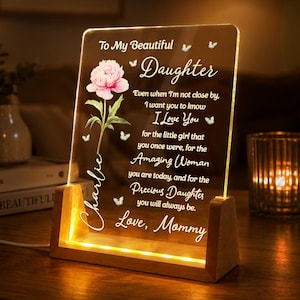 Puede incluir: Una placa rectangular de acrílico transparente sobre una base de madera iluminada, con una ilustración de peonía rosa, pequeñas mariposas y el texto: To My Beautiful Daughter, Even when I'm not close by, I want you to know I Love You, Charlie, Love, Mommy.
