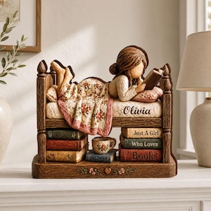 Op de afbeelding: Een decoratief beeldje van een persoon die op een bed ligt bovenop een stapel boeken. Het bedframe is van bruin hout, de persoon draagt een deken met bloemenmotief en de boeken dragen de teksten "Just A Girl Who Loves Books" en "Olivia".