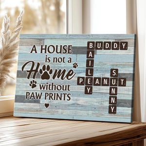 May include: A rectangular canvas print with a light blue and grey wood grain background. The text reads "A HOUSE is not a HOME without PAW PRINTS." Scrabble tiles spell out "BUDDY, ALI, PEANUT, SNY." A heart and paw prints are also visible.