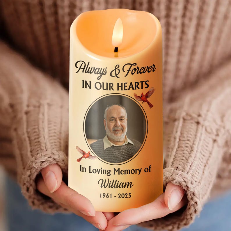 Puede incluir: Una vela LED beige con una llama parpadeante. La vela presenta el texto "Always & Forever IN OUR HEARTS" sobre una foto circular de un hombre. Debajo de la foto, se lee "In Loving Memory of William 1961-2025". Tambi&eacute;n se representan dos p&aacute;jaros rojos.