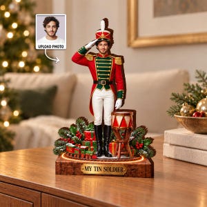 Può includere: Una statuetta personalizzata di soldatino con volto umano, uniforme rossa e verde con dettagli dorati, cappello bianco e stivali neri. La base include un trenino giocattolo, regali, rami di pino e una targa con la scritta MY TIN SOLDIER.