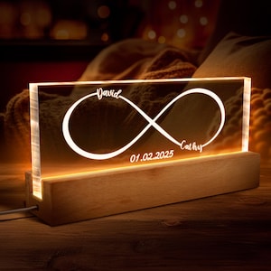 May include: A wooden base with a clear acrylic panel featuring an infinity symbol with the names "David" and "Cathy" and the date "01.02.2025" engraved. The acrylic panel is illuminated from behind, creating a warm glow.
