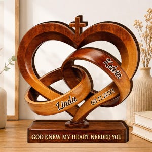 May include: A brown wooden sculpture featuring two interlocking heart shapes with a cross at the top. The piece is engraved with the names Linda and Kelvin, the date 03.11.2021, and the text GOD KNEW MY HEART NEEDED YOU on the rectangular base.