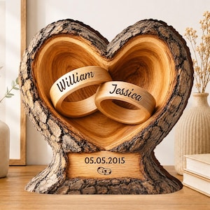 Könnte beinhalten: Ein herzförmiger Ringhalter aus Holz mit rauen Rindenkanten. Im Inneren befinden sich zwei Holzringe, die mit den Namen William und Jessica graviert sind. Ein rechteckiger Sockel zeigt das Datum 05.05.2015 und zwei ineinander verschlungene Ringe.