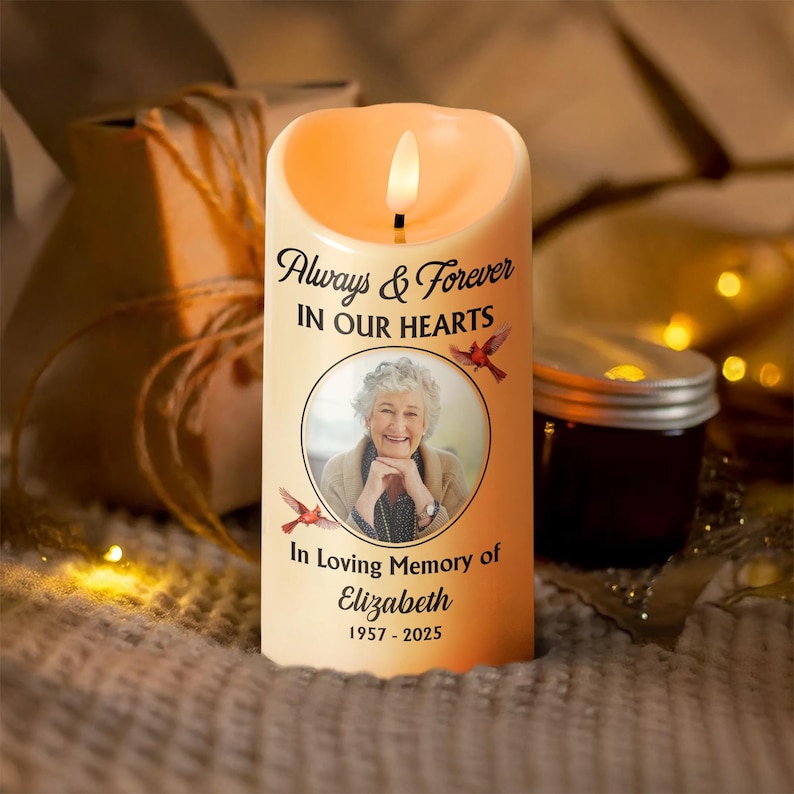 Puede incluir: Una vela LED color crema con un brillo c&aacute;lido, con el texto "Always & Forever IN OUR HEARTS" y una foto de una mujer. La vela incluye el texto "In Loving Memory of Elizabeth 1957-2025" y dos ilustraciones de p&aacute;jaros rojos.