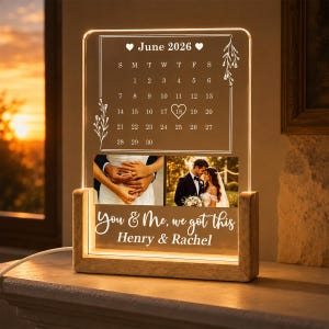 Op de afbeelding: Een rechthoekige acrylplaat met houten voetstuk met een verlichte kalender van juni 2026 met een hartje rond de 18e, twee afgedrukte foto's van een koppel en de tekst "You & Me, we got this" boven de namen "Henry & Rachel".