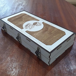 Könnte beinhalten: Eine weiße Holzkiste mit einer braunen Holzeinlage und einem Design aus zwei Würfeln. Die Kiste hat zwei silberne Verschlüsse.