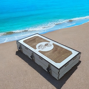 Könnte beinhalten: Rechteckige Holzkiste mit hellbrauner Oberseite und weißen Akzenten. Die Oberseite hat ein kreisförmiges Design mit Würfeln. Die Kiste befindet sich an einem Sandstrand mit dem Meer im Hintergrund.
