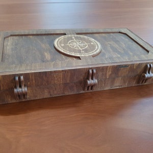 Puede incluir: Caja rectangular de madera marrón oscuro. La tapa presenta un diseño de rosa de los vientos. La caja tiene cierres decorativos y veta de madera visible, creando una estética rústica. La caja está cerrada.