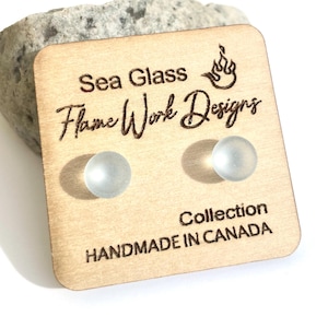 May include: Sea Glass Flame Work Designs stud earrings on a wooden card. The card has the text "Sea Glass Flame Work Designs Collection Handmade in Canada". The earrings are small, round, and frosted, with a light blue hue.
