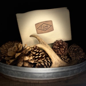 May include: A white canvas zippered pouch with a brown leather patch that says "J Woods Enterprises". The pouch is sitting in a metal bowl with pine cones and a deer antler.
