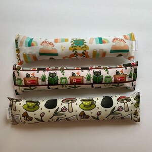 May include: Three cat toys with colourful patterns. The top toy has rainbows, mushrooms, and books on a white background. The middle toy features black cats reading books. The bottom toy has mushrooms, teacups, and books.