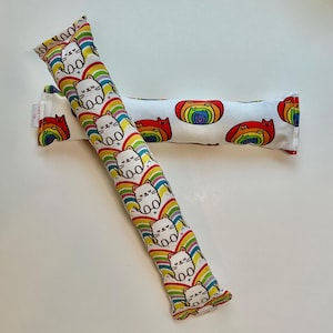 May include: Two cat toys. One is a long, rectangular shape with a repeating pattern of white cats on rainbows. The other is a similar shape with a repeating pattern of rainbow cats. Both toys are on a white surface.