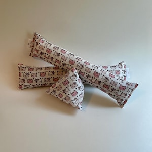 May include: Four catnip toys in various shapes. The toys are white with a repeating pattern of pink and brown cat faces. Two are long and rectangular, one is small and rectangular, and one is triangular.