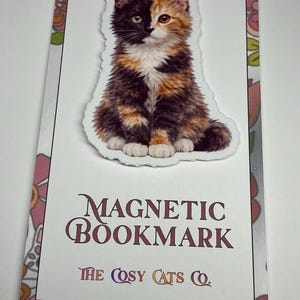 May include: A magnetic bookmark featuring a fluffy calico cat illustration. The cat has a split face of black and orange fur. The bookmark has the words "MAGNETIC BOOKMARK" and "THE COSY CATS CO." printed on it, with a floral border.