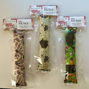 May include: Three cat toys in clear packaging. Each toy is a long, rectangular shape with a different patterned fabric. The patterns include flowers, daisies, and a garden scene with black cat illustrations. Each package has a label that reads "Kitty Kicker".