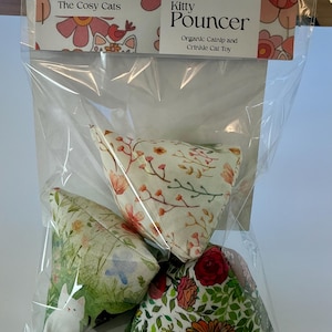Può includere: Un sacchetto di plastica trasparente contiene tre giocattoli triangolari per gatti. I giocattoli sono ricoperti da motivi floreali nei colori rosa, rosso, verde e bianco. L'etichetta in alto dice "The Cosy Cats" e "Kitty Pouncer".