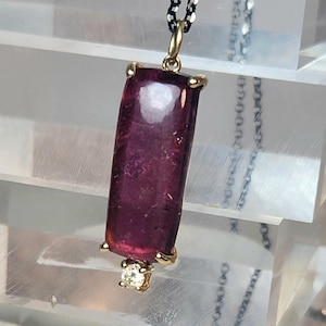 May include: A pendant necklace featuring a rectangular, deep red gemstone. The gemstone is set in a gold-colored bezel with a small diamond accent at the bottom. The chain is a dark, textured metal.