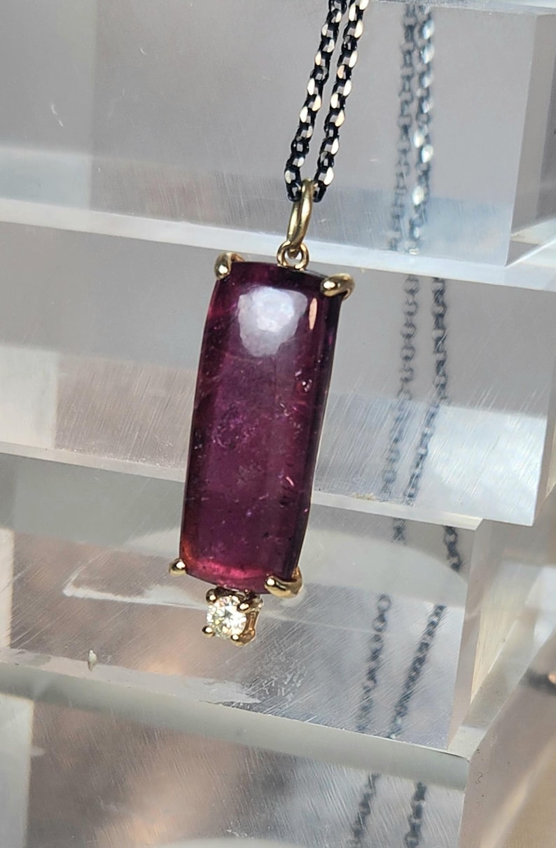 May include: A pendant necklace featuring a rectangular, deep red gemstone. The gemstone is set in a gold-colored bezel with a small diamond accent at the bottom. The chain is a dark, textured metal.