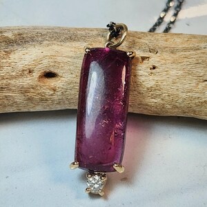 May include: A rectangular, deep magenta pendant with a gold setting and a small diamond accent. The pendant hangs from a delicate, dark chain. The gemstone is approximately 1 inch in length.