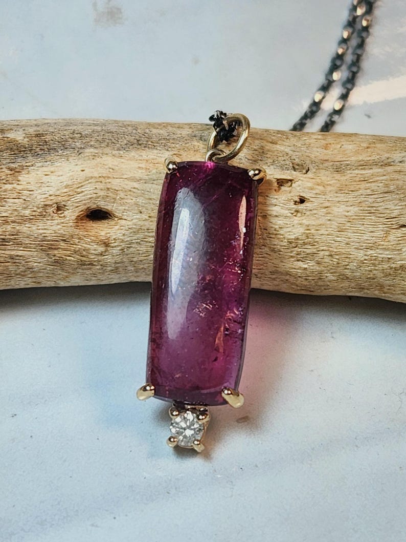 May include: A rectangular, deep magenta pendant with a gold setting and a small diamond accent. The pendant hangs from a delicate, dark chain. The gemstone is approximately 1 inch in length.