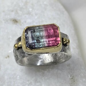 May include: A silver ring featuring a rectangular gemstone with pink, green, and blue hues, set in a gold bezel. The ring band has a textured finish and small gold accents. The ring is a piece of jewellery.