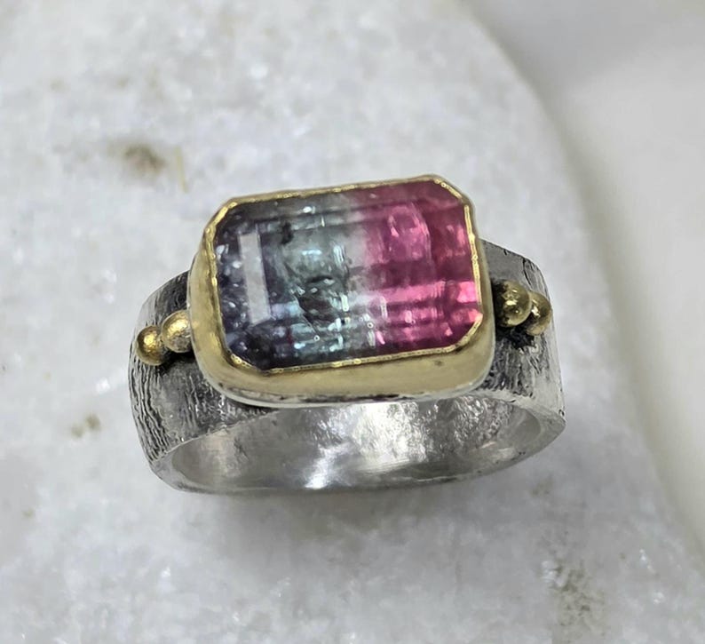 May include: A silver ring featuring a rectangular gemstone with pink, green, and blue hues, set in a gold bezel. The ring band has a textured finish and small gold accents. The ring is a piece of jewellery.