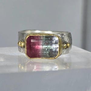 May include: A silver ring featuring a rectangular, bi-colour gemstone. The gemstone, framed in gold, displays a gradient of pink and green hues. The ring band has a textured finish and small gold accents.
