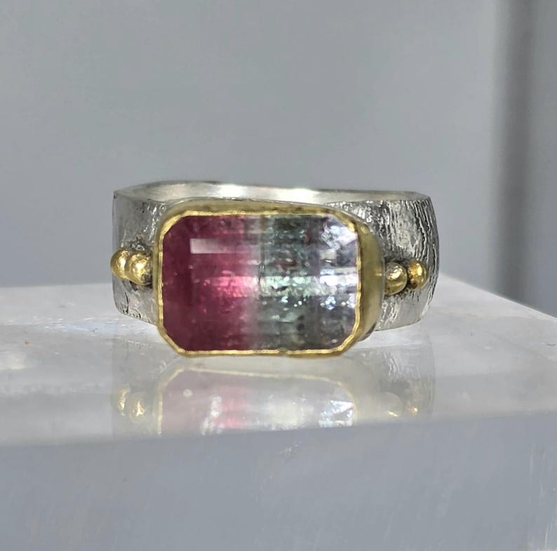 May include: A silver ring featuring a rectangular, bi-colour gemstone. The gemstone, framed in gold, displays a gradient of pink and green hues. The ring band has a textured finish and small gold accents.