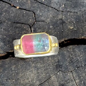 May include: A silver ring with a rectangular gemstone displaying pink, green, and blue colours. The gemstone is set in a gold bezel. The ring has a textured band and small gold accents on each side of the gemstone.