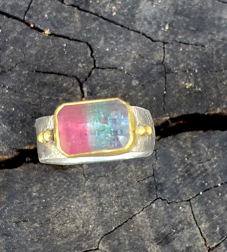 May include: A silver ring with a rectangular gemstone displaying pink, green, and blue colours. The gemstone is set in a gold bezel. The ring has a textured band and small gold accents on each side of the gemstone.
