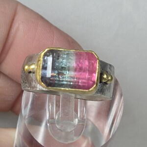 May include: A silver ring featuring a rectangular watermelon tourmaline gemstone. The gemstone is framed in gold and displays pink, green, and clear sections. Small gold accents are on either side of the stone.