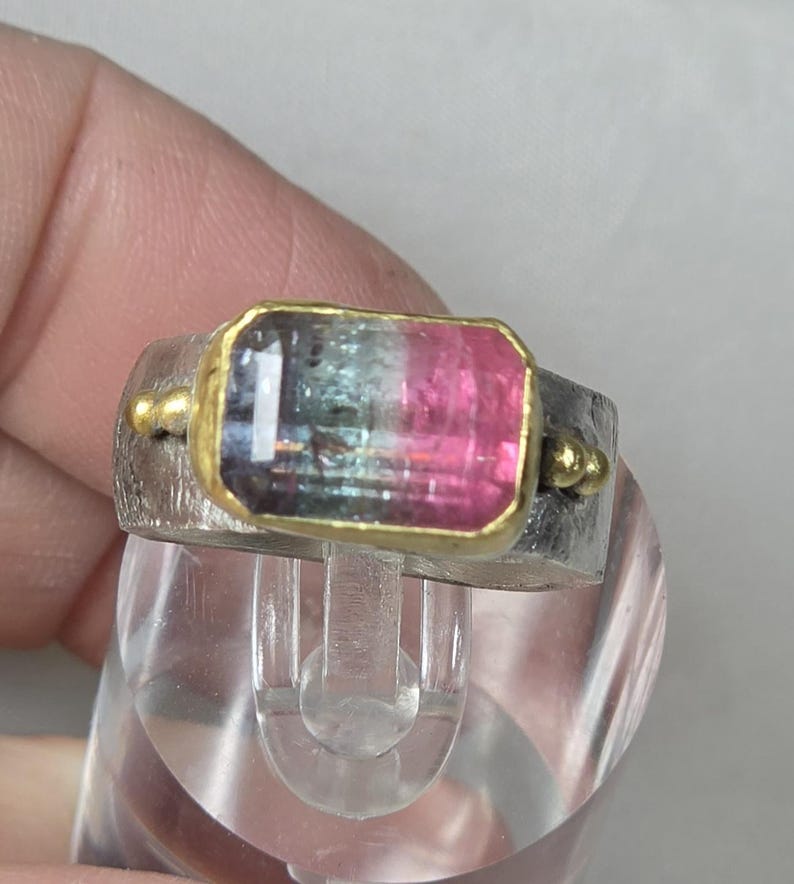 May include: A silver ring featuring a rectangular watermelon tourmaline gemstone. The gemstone is framed in gold and displays pink, green, and clear sections. Small gold accents are on either side of the stone.