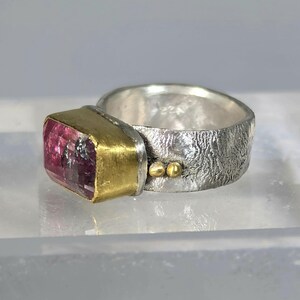 May include: A silver ring with a textured band and a rectangular pink gemstone set in a gold bezel. Two small gold beads accent the band. The ring is a piece of jewellery.