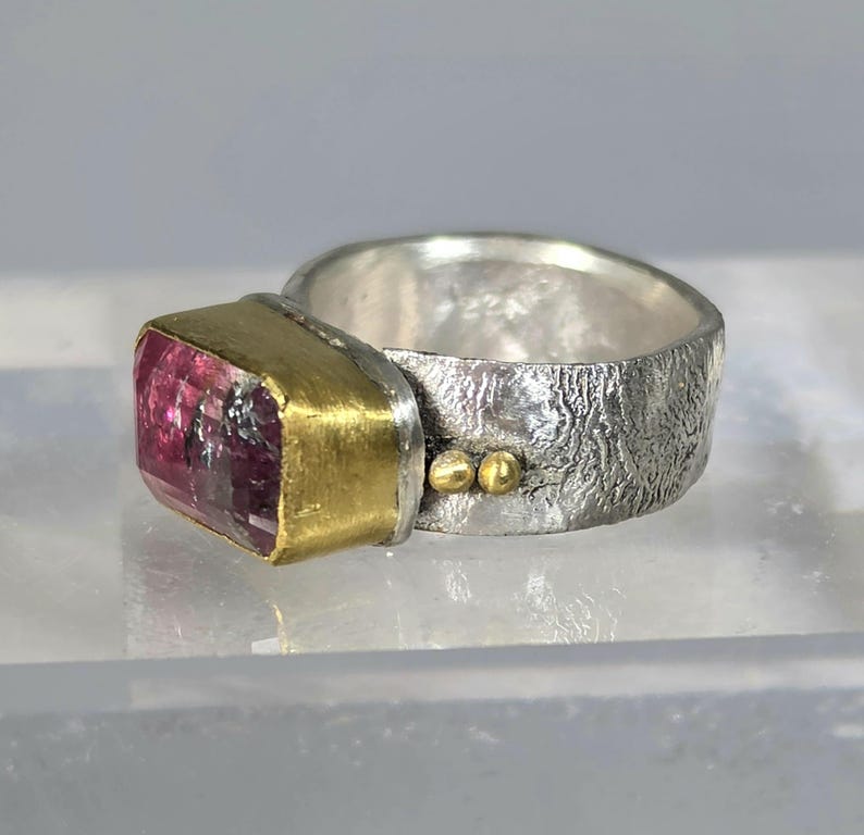 May include: A silver ring with a textured band and a rectangular pink gemstone set in a gold bezel. Two small gold beads accent the band. The ring is a piece of jewellery.