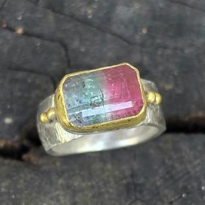 May include: A silver ring featuring a rectangular watermelon tourmaline gemstone. The gemstone displays pink and green hues, set in a gold bezel. The ring has a textured band and gold detailing.