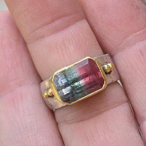 May include: A silver ring featuring a rectangular watermelon tourmaline gemstone. The gemstone is framed in gold and shows a gradient of green, pink, and clear hues. The ring has a textured band and small gold accents on either side of the gemstone.