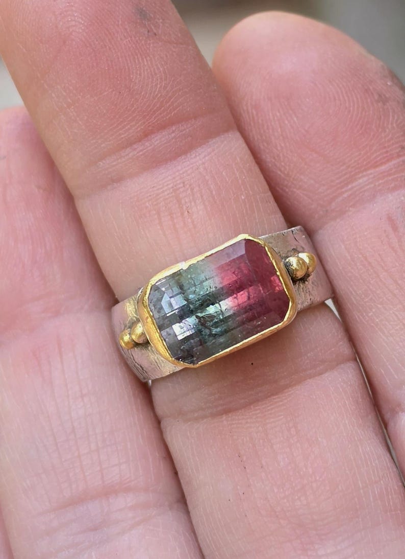 May include: A silver ring featuring a rectangular watermelon tourmaline gemstone. The gemstone is framed in gold and shows a gradient of green, pink, and clear hues. The ring has a textured band and small gold accents on either side of the gemstone.