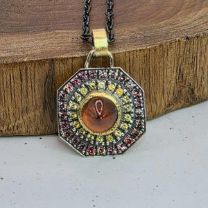 Puede incluir: Collar con colgante plateado con diseño geométrico. El colgante presenta una piedra central de color ámbar rodeada de piedras preciosas amarillas y rosas. El collar tiene una cadena oscura y una fianza dorada.