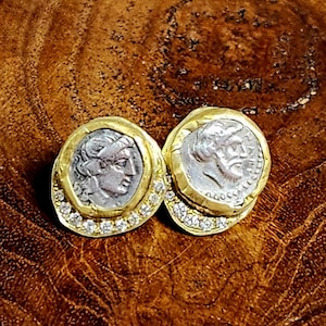 May include: A pair of gold earrings with a silver coin design. Each earring features a profile of a person's face on the coin, surrounded by a circle of small diamonds.