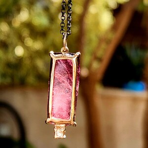 May include: A pendant necklace featuring a rectangular pink gemstone set in a gold-colored frame. The gemstone has internal veining. The pendant hangs from a dark chain. A small, clear stone is set at the bottom of the pendant.