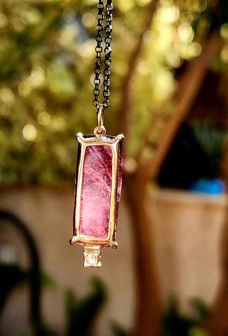 May include: A pendant necklace featuring a rectangular pink gemstone set in a gold-colored frame. The gemstone has internal veining. The pendant hangs from a dark chain. A small, clear stone is set at the bottom of the pendant.