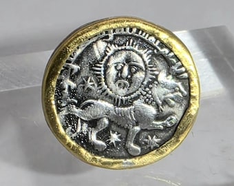 May include: A circular coin-shaped pendant featuring a silver-toned relief of a lion walking beneath a sun face and stars, framed by a thick, textured gold-toned border. The central design is oxidized to highlight the detailed engravings.