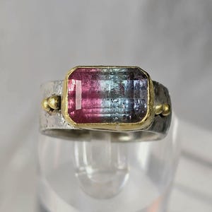 May include: A silver ring featuring a rectangular watermelon tourmaline gemstone. The gemstone is framed in gold and displays pink, blue, and green colours. The ring has a textured band with gold accents on either side of the gemstone.