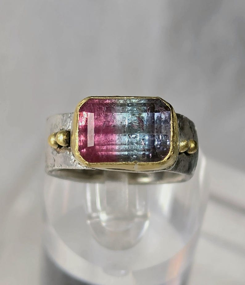 May include: A silver ring featuring a rectangular watermelon tourmaline gemstone. The gemstone is framed in gold and displays pink, blue, and green colours. The ring has a textured band with gold accents on either side of the gemstone.