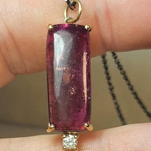 May include: A pendant necklace featuring a rectangular, deep pink gemstone with gold accents. A small, round diamond is set at the bottom. The pendant hangs from a delicate, dark chain, creating an elegant jewelry piece.