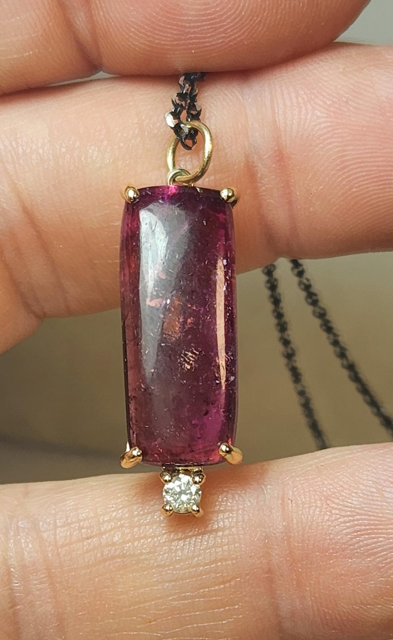 May include: A pendant necklace featuring a rectangular, deep pink gemstone with gold accents. A small, round diamond is set at the bottom. The pendant hangs from a delicate, dark chain, creating an elegant jewelry piece.