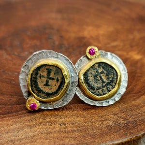 May include: A pair of stud earrings featuring ancient coin designs. Each earring has a silver base with a gold-colored bezel surrounding a dark coin. A small, pink gemstone is set on each earring. The earrings are displayed on a wooden surface.